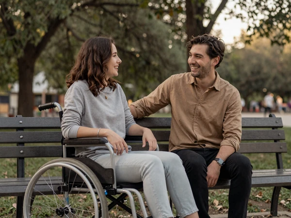 Two friends laughing together on a park bench, one a wheelchair user, in soft afternoon light